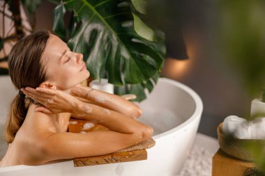 Küvetin kenarına yaslanmış, banyoda tropik bitkilerle süslenmiş huzurlu genç bir kadın.