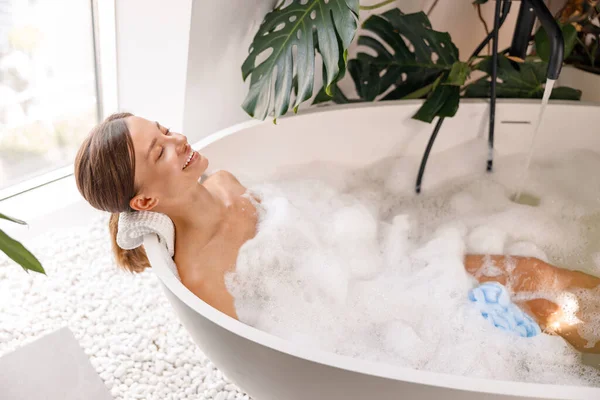 Donna rilassata cercando felice mentre prende il bagno di bolla nel suo bel bagno decorato con piante — Foto Stock