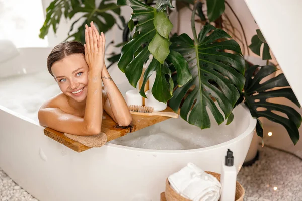Ritratto di giovane donna felice con un bel sorriso guardando la macchina fotografica mentre si appoggia sul lato del bagno bianco decorato con piante tropicali — Foto Stock