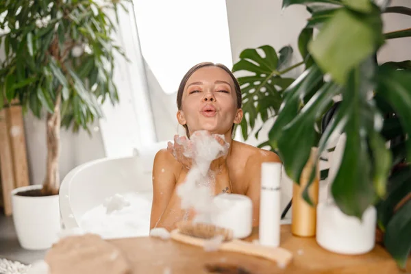 Lekfull ung kvinna blåser tvålskum medan du tar bubbelbad på spa resort — Stockfoto