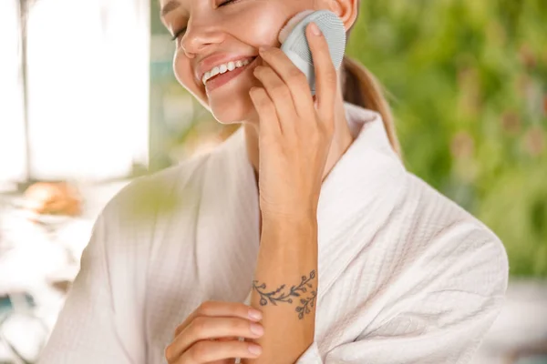 Retrato recortado de una mujer joven que usa albornoz sonriendo mientras usa un cepillo facial de silicona para limpiar la piel — Foto de Stock