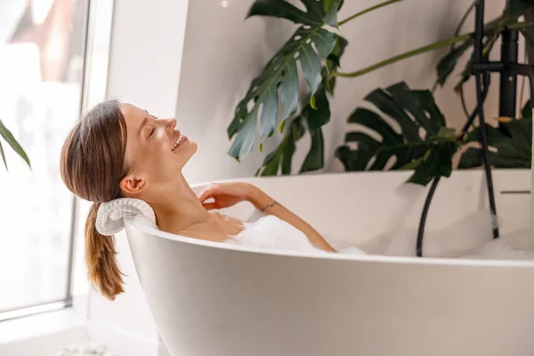 熱帯植物で飾られたバスルームで泡風呂に入りながら笑顔明るい若い女性の側面図 — ストック写真