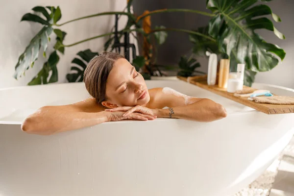 Giovane donna sognante appoggiata su un lato vasca da bagno, rilassante con gli occhi chiusi a casa — Foto Stock