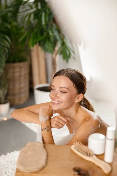 Jovencita alegre sonriendo y apoyada en la bañera mientras se baña en el balneario — Foto de Stock