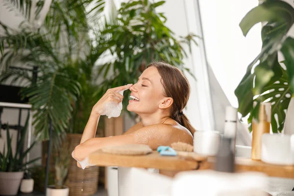 Hoş genç bir kadın eğleniyor. Tropik spa merkezinde banyo yaparken sabun köpüğüyle oynuyor.