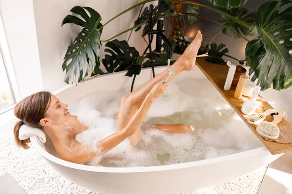 Radosna młoda kobieta leżąca w wannie i goląca nogi jednorazową golarką. Kosmetyki do pielęgnacji ciała na drewnianej półce nad wanną — Zdjęcie stockowe