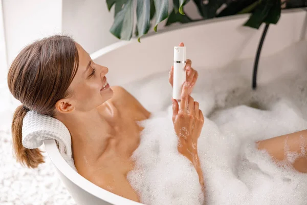 Joven alegre sosteniendo el producto de cuidado corporal mientras se baña en la bañera con espuma de burbuja — Foto de Stock