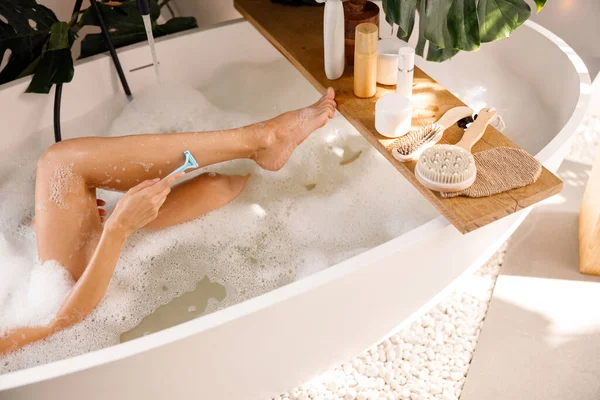 Nogi młodej kobiety golącej się brzytwą do golenia podczas kąpieli. Kosmetyki do pielęgnacji ciała na drewnianej półce nad wanną — Zdjęcie stockowe