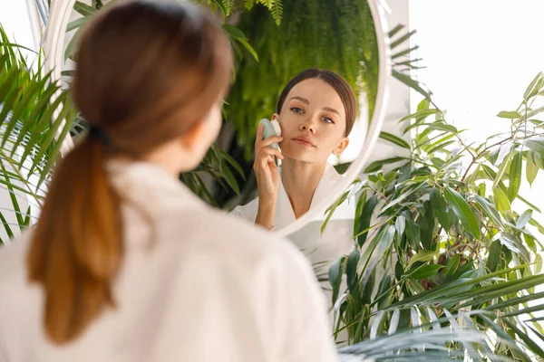 Молодая женщина в халате смотрит на себя в зеркало, используя силиконовую щетку для лица, чтобы очистить кожу, стоя в ванной комнате, украшенной зелеными растениями — стоковое фото