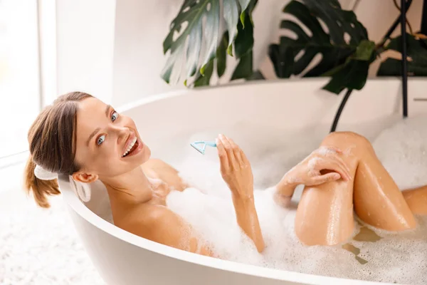 Feliz joven mujer sonriendo a la cámara, sosteniendo afeitadora desechable para afeitarse las piernas en el baño con espuma —  Fotos de Stock