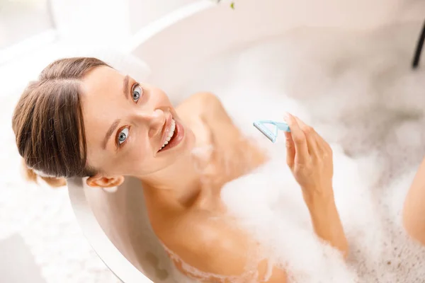 Feliz joven mujer sonriendo a la cámara, sosteniendo afeitadora desechable para afeitarse las piernas en el baño con espuma —  Fotos de Stock