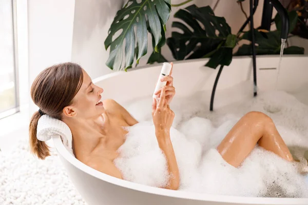 Mujer joven feliz sonriendo mientras toma un baño de burbujas y cuida su piel en un spa de lujo —  Fotos de Stock