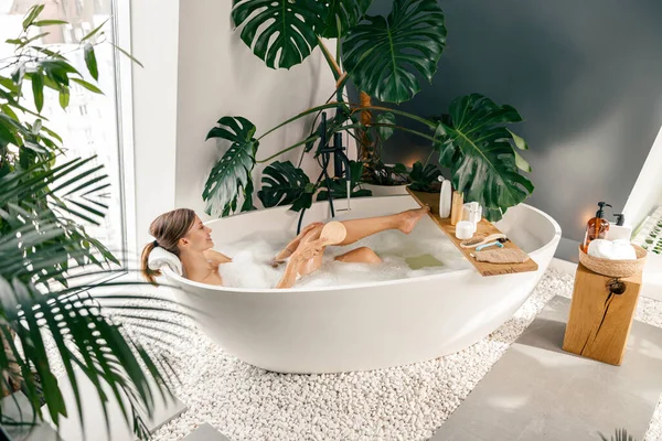 Ontspannen jonge vrouw met behulp van borstel, terwijl het nemen van bubbelbad in moderne badkamer versierd met tropische planten — Stockfoto