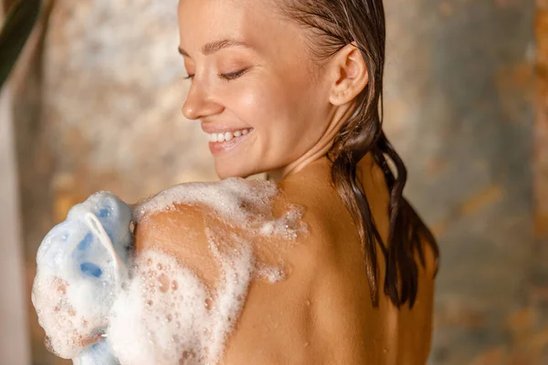 Vacker ung kvinna tillämpa dusch gel med hjälp av loofah svamp medan du tar dusch — Stockfoto