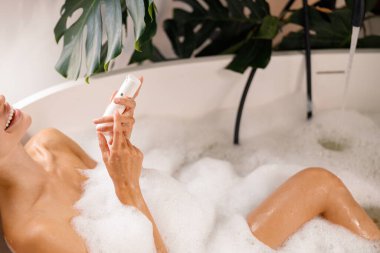 Rahatlamış genç bir kadın köpük banyosu yaparken ve vücut bakım ürünlerini tutarken, lüks bir spa merkezinde kendine bakarken gülümsüyor.