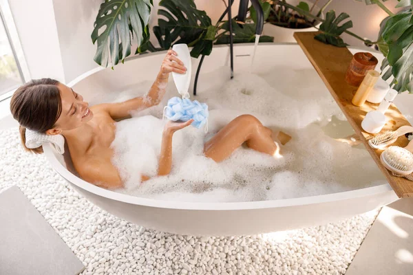 Relajada joven mujer verter gel de ducha de botella sobre esponja de esponja de esponja mientras se baña —  Fotos de Stock