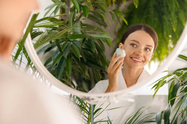 Портрет молодой женщины, радостно смотрящей на себя в зеркало, очищающей кожу силиконовой кисточкой в ванной комнате, украшенной зелеными растениями — стоковое фото