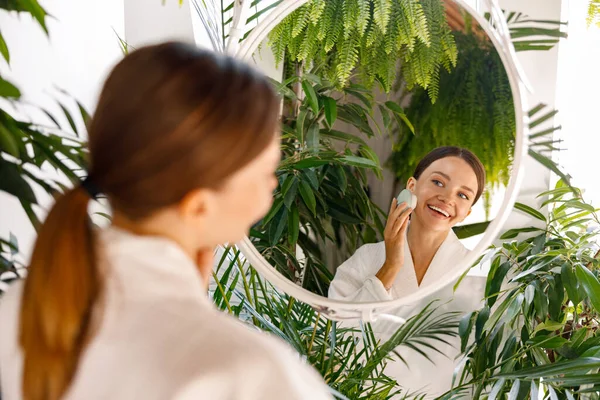 Счастливая молодая женщина в халате наслаждается процедурой очищения кожи силиконовой щеткой для лица, стоя в ванной комнате, украшенной зелеными растениями — стоковое фото