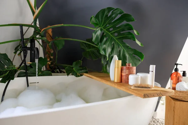 Mensola in legno per prodotti di bellezza e cura del corpo su mensola in legno sopra moderna vasca da bagno riempita di bolle bianche. Accogliente elegante bagno decorato con verde pianta tropicale Monstera — Foto Stock