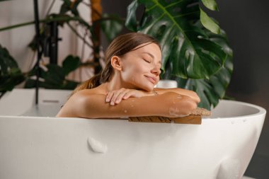 Küvetin kenarına yaslanmış, tropikal bitkilerle süslenmiş banyoda dinlenen hayalperest genç kadın.