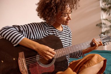 Gülümseyen genç bir kadın evde gitar çalıyor.