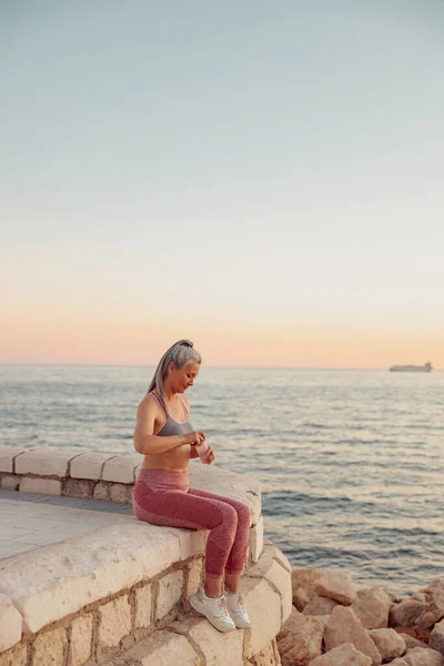 Mujer deportiva sentada en el borde del terraplén, sosteniendo una botella de agua —  Fotos de Stock