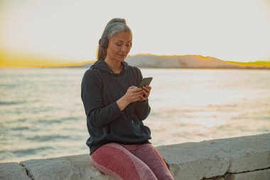 Barajda oturan, kulaklık takan ve telefona bakan bir kadın.