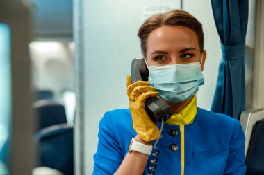 Tıbbi maskeli kadın hostes uçakta telefonla konuşuyor.