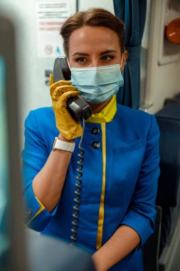 Tıbbi maskeli uçuş görevlisi uçakta telefonla konuşuyor.