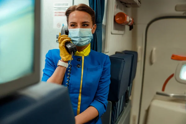 Letuška v lékařské masce telefonicky v kabině letadla — Stock fotografie