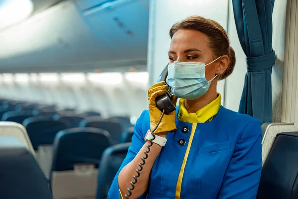Letuška v lékařské masce mluví po telefonu v kabině letadla — Stock fotografie