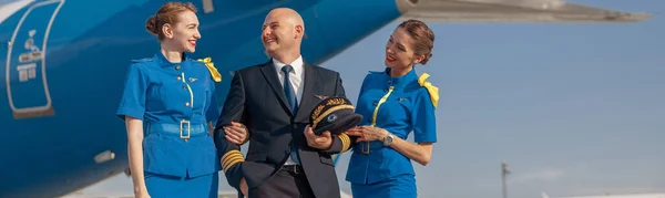 Возбужденный пилот с двумя привлекательными стюардессами, стоящими вместе перед самолетом и улыбающимися после посадки — стоковое фото