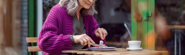Mooie oudere dame in paars gebreide jas eet heerlijk dessert aan tafel op terras cafe buiten — Stockfoto