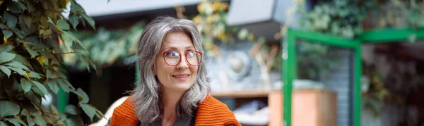 Mujer madura feliz con gafas come postre sentado en la pequeña mesa al aire libre —  Fotos de Stock