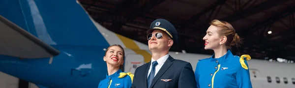 Piloto feliz em uniforme e óculos de sol aviador andando junto com duas aeromoças em uniforme azul na frente de grande avião de passageiros no hangar do aeroporto — Fotografia de Stock