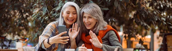 Mulheres maduras positivas amigos acenam as mãos no videochat via smartphone no café de rua — Fotografia de Stock