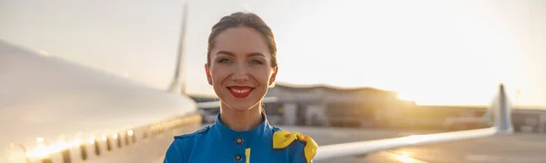 Retrato de bela aeromoça com lábios vermelhos em uniforme azul sorrindo para a câmera, posando ao ar livre com avião comercial perto do terminal em um aeroporto no fundo — Fotografia de Stock