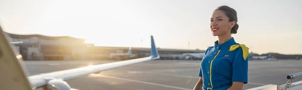 Όμορφη αεροσυνοδός με μπλε στολή να χαμογελάει, να στέκεται έξω στο ηλιοβασίλεμα. Εμπορικό αεροπλάνο κοντά στον τερματικό σταθμό σε ένα αεροδρόμιο στο παρασκήνιο — Φωτογραφία Αρχείου