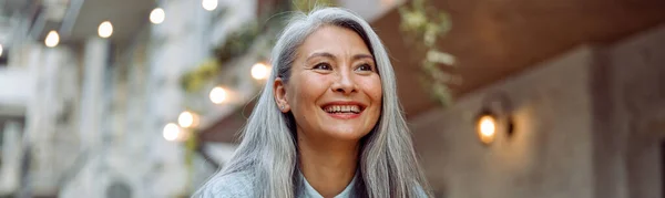 Cheerful mature Asian woman with glass of water and phone sits at small table outdoors — Stock Photo, Image