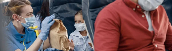 Nettes Mädchen mit schützender Gesichtsmaske, das auf eine Stewardess blickt, die kleinen Passagieren an Bord Mittagessen serviert. Flugreisen während der Covid19-Pandemie — Stockfoto