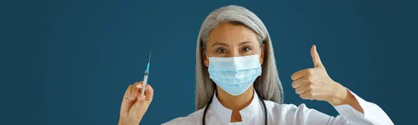 Ώριμη Ασιάτισσα γιατρός με μάσκα κρατάει τη σύριγγα και δείχνει τον αντίχειρα πάνω σε μπλε φόντο — Φωτογραφία Αρχείου