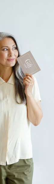 Thoughtful hoary haired Asian lady customer holds gift card standing on light grey background — Stock Photo, Image