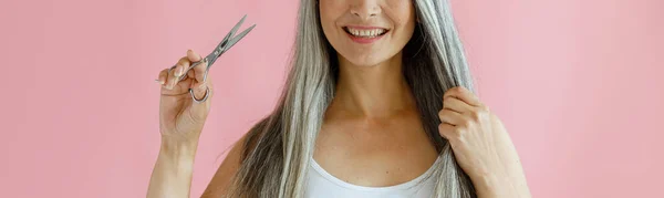 Веселая азиатка держит прядь серебряных волос и ножниц на розовом фоне — стоковое фото