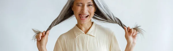 Idade média asiático senhora grimaces e detém longos cabelos prateados no fundo cinza claro — Fotografia de Stock