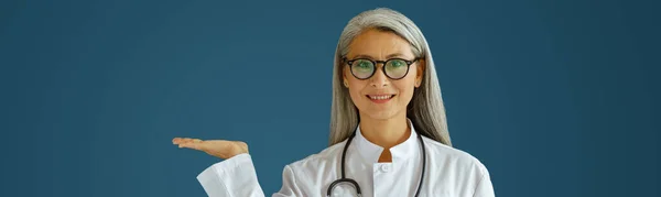 Middelbare leeftijd vrouwelijke arts met bril en stethoscoop punten opzij op blauwe achtergrond in de studio — Stockfoto