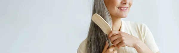 Nachdenkliche Asiatin mittleren Alters bürstet graue Haare auf hellem Hintergrund — Stockfoto