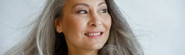 Happy middle aged Asian woman with flying grey locks stands on light background — Stock Photo, Image