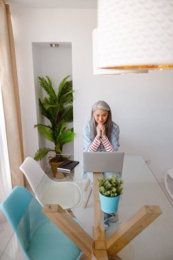 Masada oturan ve evde dizüstü bilgisayarla çalışan bir kadın.
