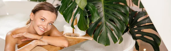 Portrait of attractive young woman smiling at camera while leaning on side of white bath decorated with tropical plant — Stock Photo, Image