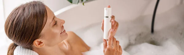 Fröhliche junge Frau hält Körperpflegemittel in der Badewanne mit Schaumstoffschaumstoff — Stockfoto